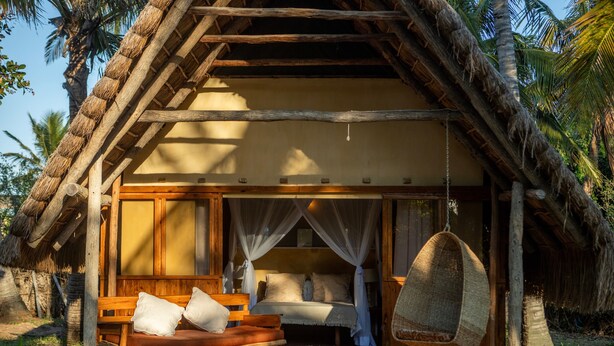A thatched-roof hut with a bed, a hanging chair, and a bench. - Turtle Cove Lodge and Yoga Shala (Tofo)