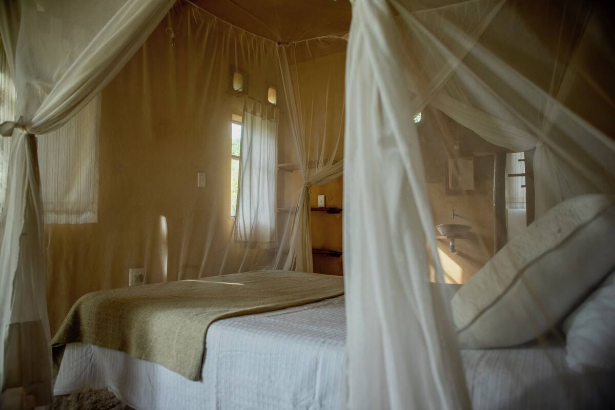 Een kamer met een bed dat overdekt is door een hemelbed, een raam met gordijnen, en een wastafel op de achtergrond.
