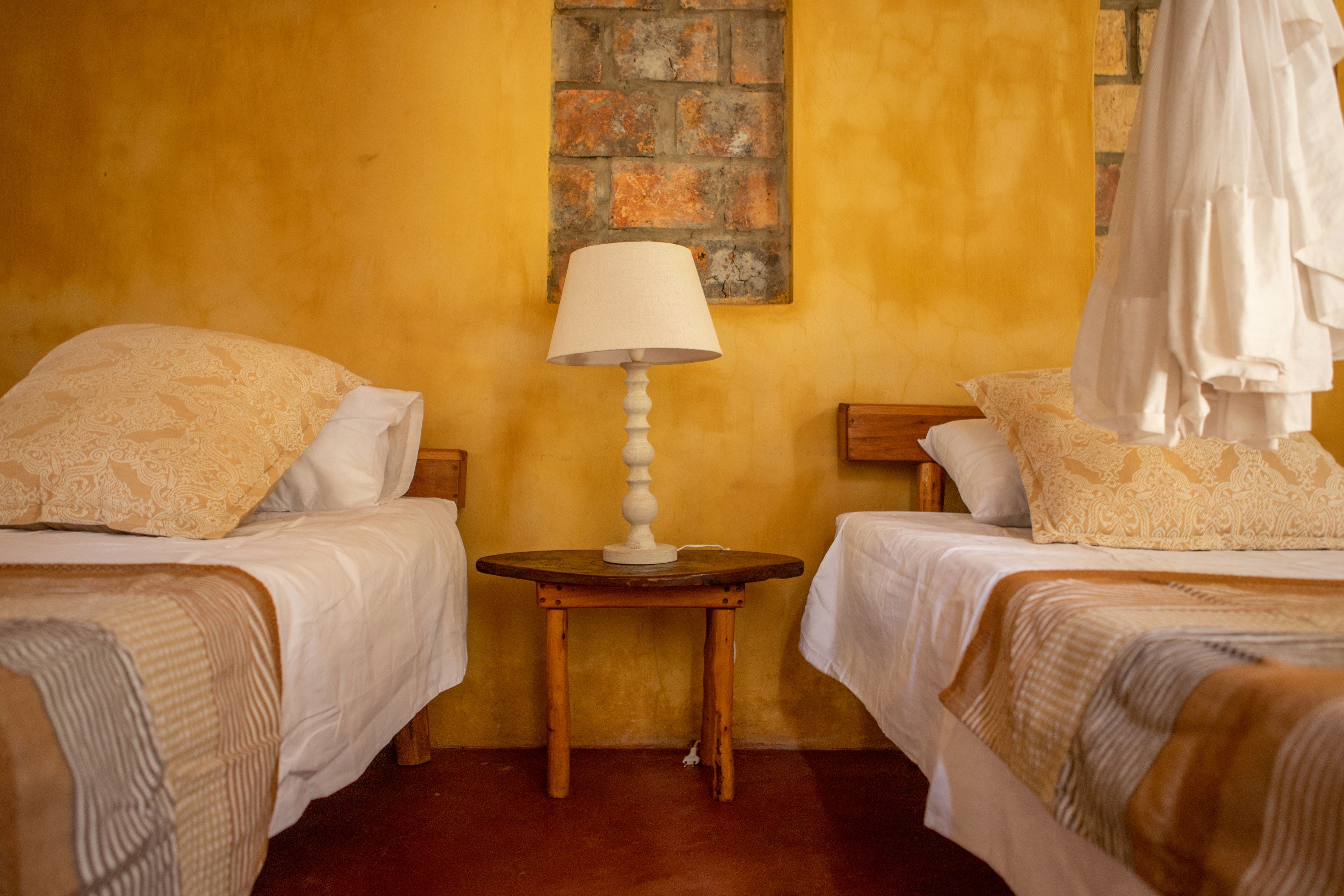 Una habitación con dos camas, una pequeña mesa de madera con una lámpara y una pared de ladrillo.
