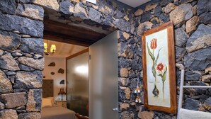 A stone-walled room with a bed, a chair, a lamp, and a framed botanical illustration on the wall.