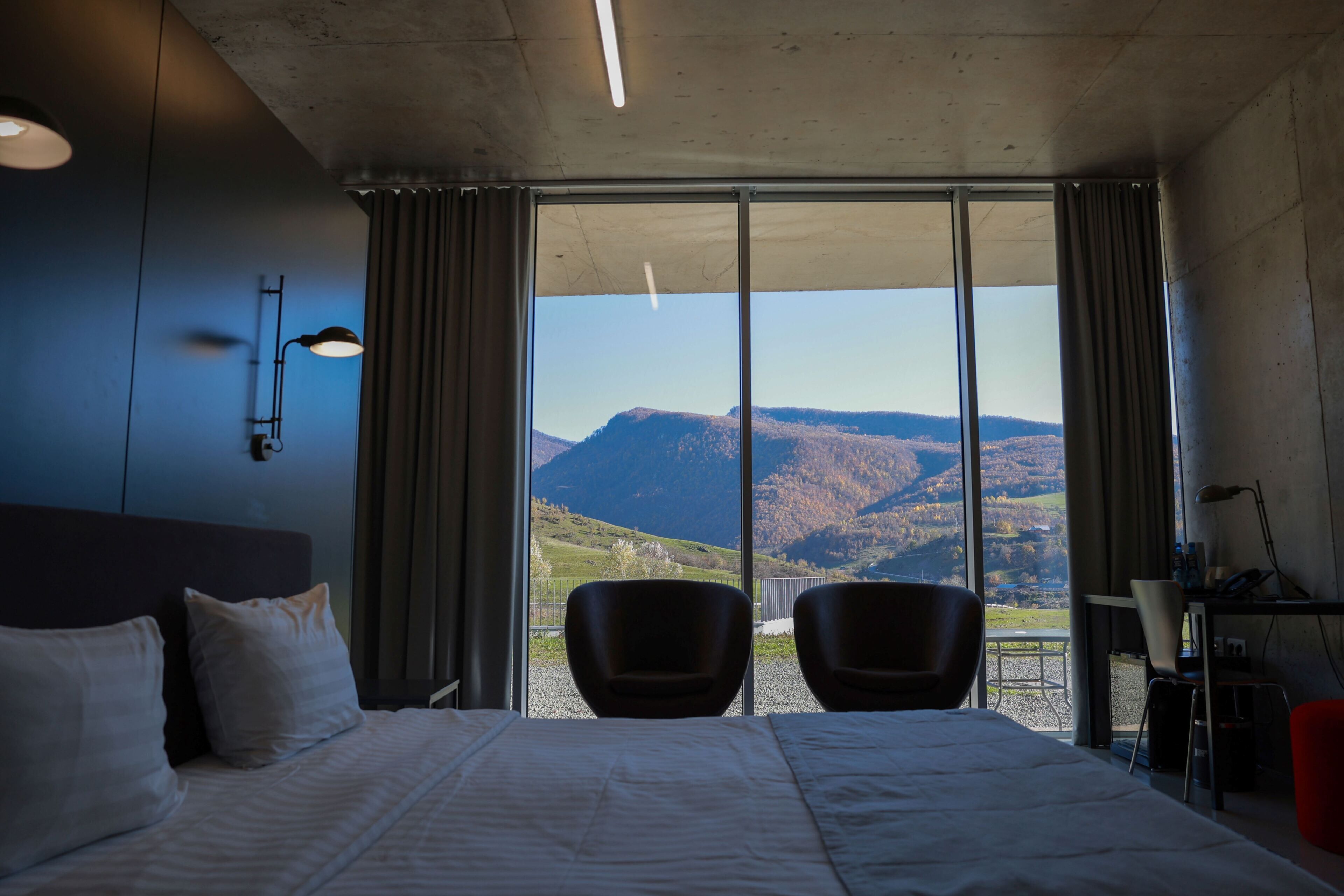 Habitación de hotel con una cama grande, dos sillas, un escritorio con una lámpara y vistas a las montañas a través de ventanales del suelo al techo.