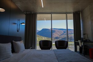 Habitación de hotel con una cama grande, dos sillas, un escritorio con una lámpara y vistas a las montañas a través de ventanales del suelo al techo.