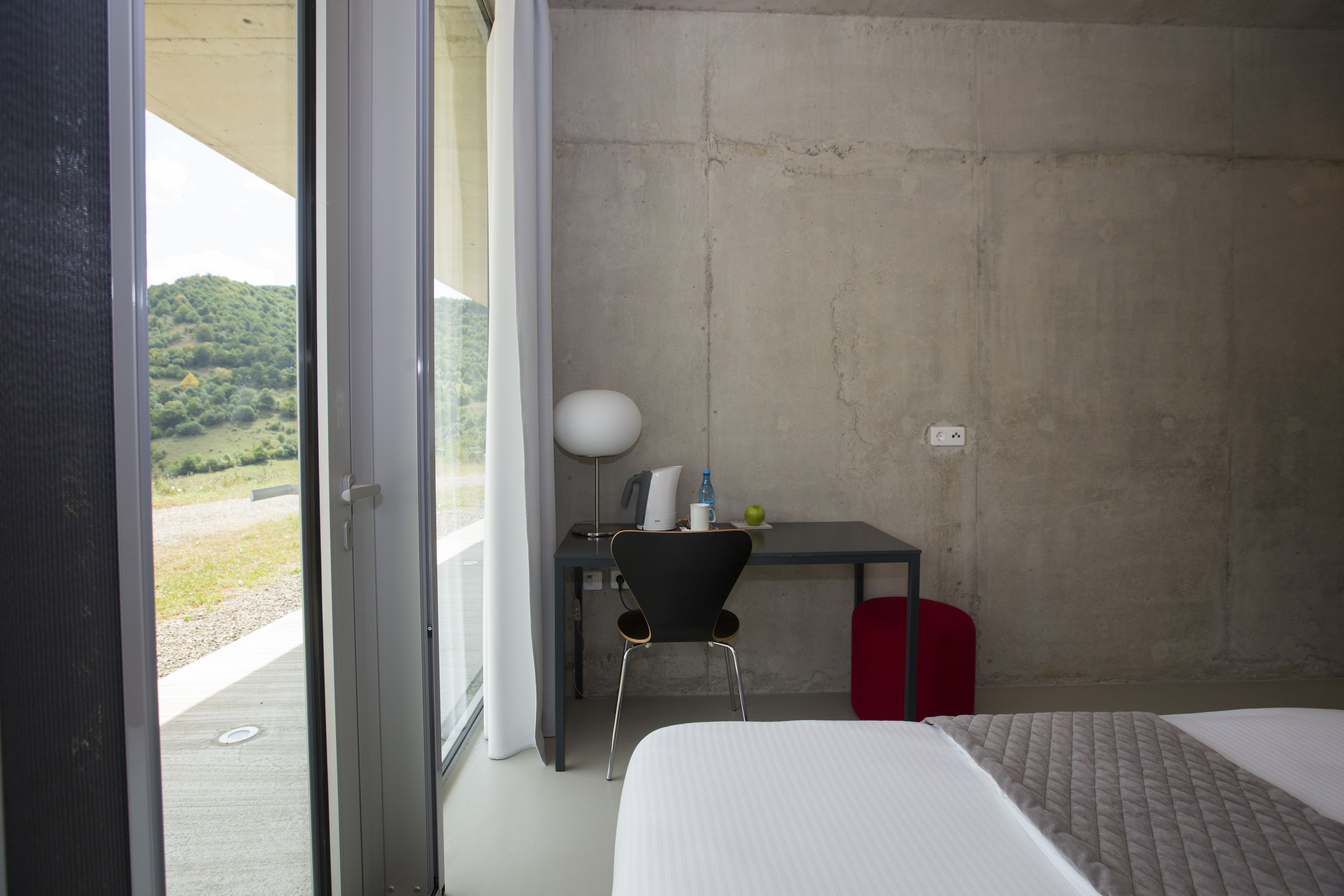 Habitación de hotel con cama, escritorio con silla, lámpara y vista a un paisaje verde a través de una puerta de cristal.