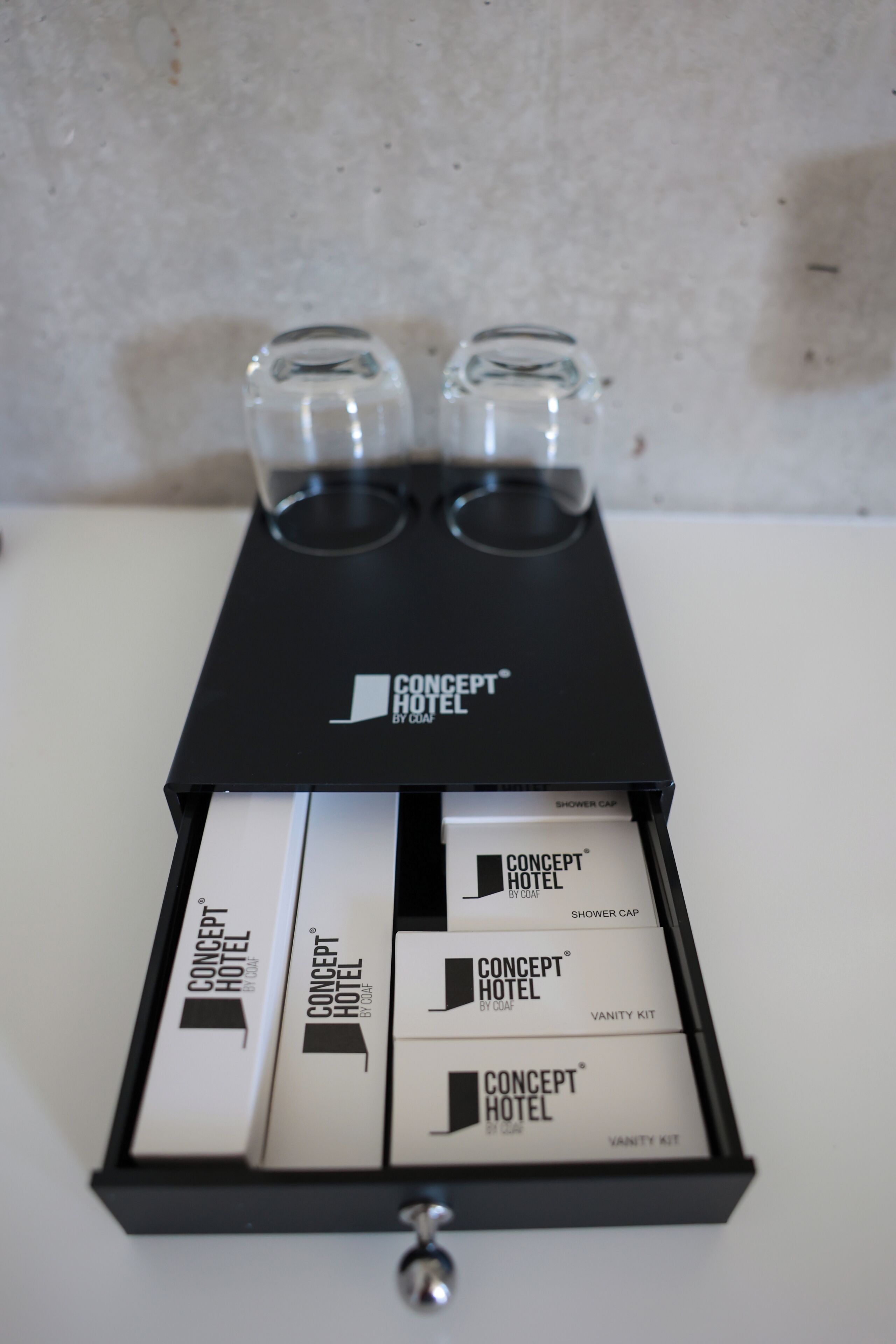 Un cajón con dos vasos y varios artículos de tocador de hotel, incluyendo una bolsa para el gorro de ducha, kit de aseo y la marca del hotel.