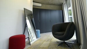 Una habitación moderna con un ventanal grande, una silla negra, un reposapiés rojo y un cuadro enmarcado apoyado contra la pared.