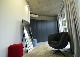 Una habitación moderna con un ventanal grande, una silla negra, un reposapiés rojo y un cuadro enmarcado apoyado contra la pared.