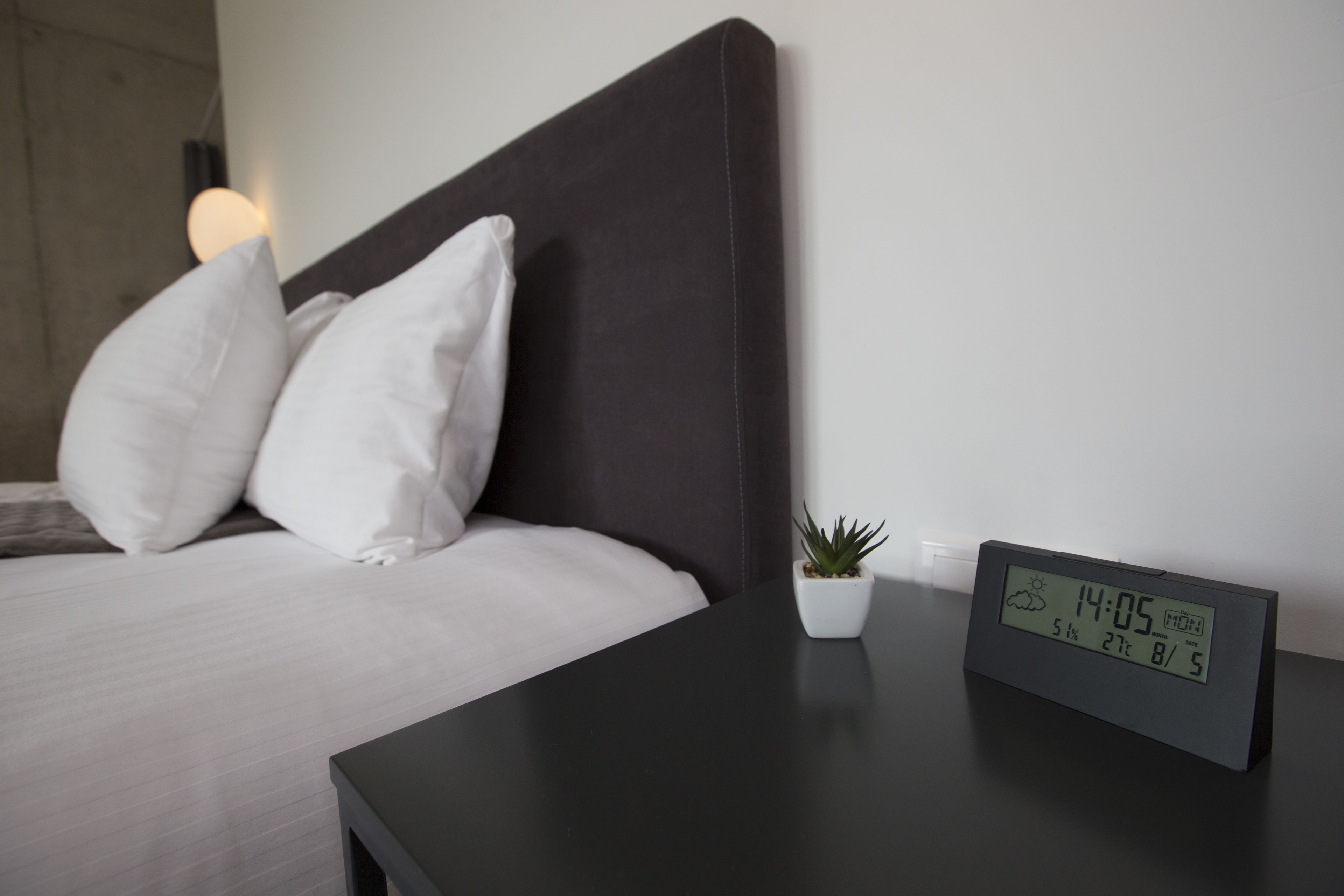A neatly made bed with white pillows and a small potted plant on a bedside table. A digital clock displays the time and weather.