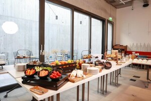 Disposición de bufé con diversos alimentos y una larga mesa en un interior moderno con ventanas amplias.