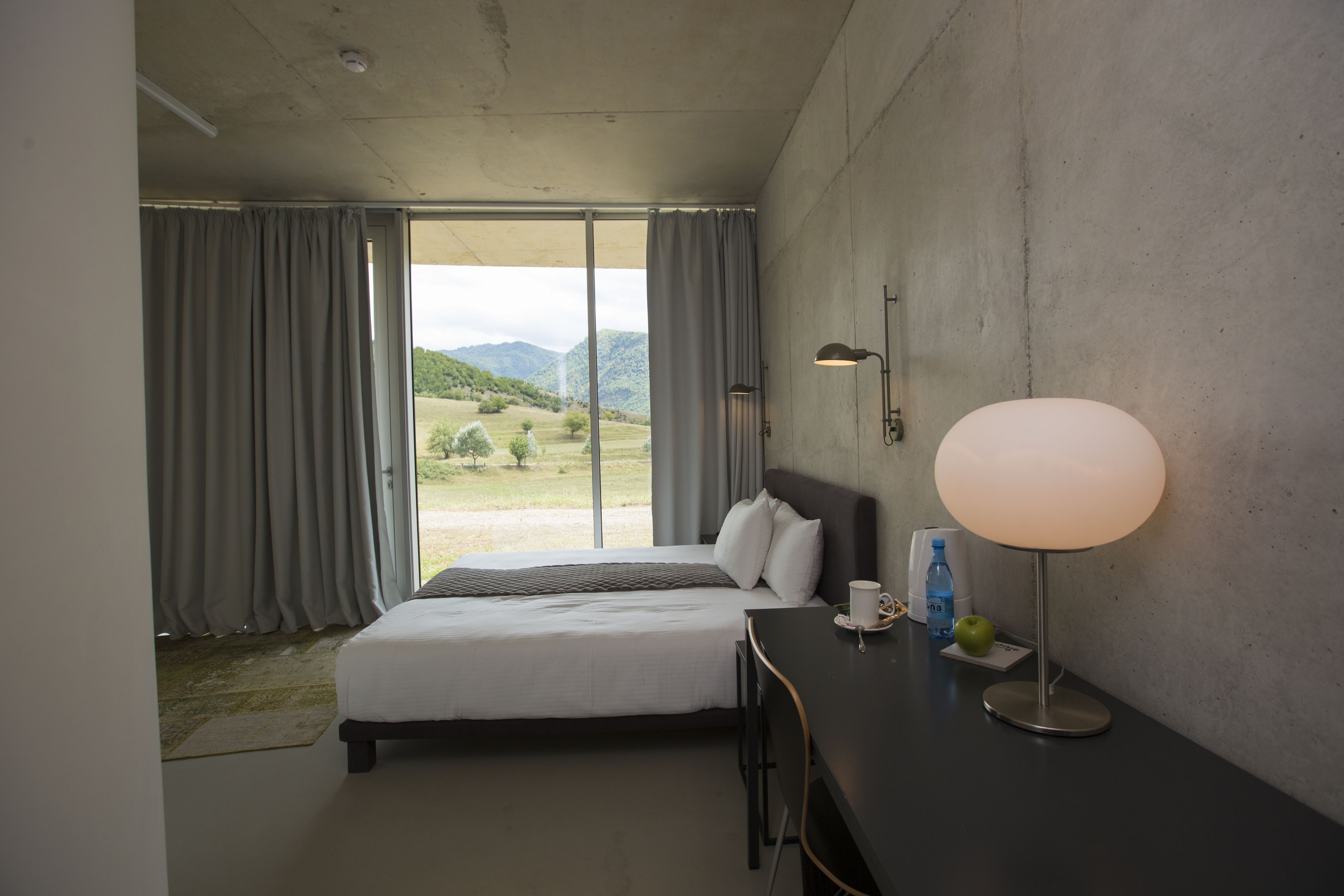 A hotel room with a bed, a desk with a lamp, a cup, and a bottle.