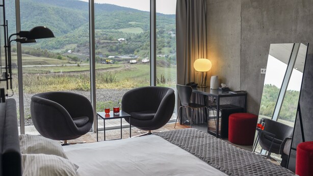 Habitación de hotel moderna con una cama grande, dos sillas negras, un escritorio con lámpara y vistas a colinas verdes.