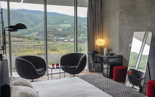 Habitación de hotel moderna con una cama grande, dos sillas negras, un escritorio con lámpara y vistas a colinas verdes.