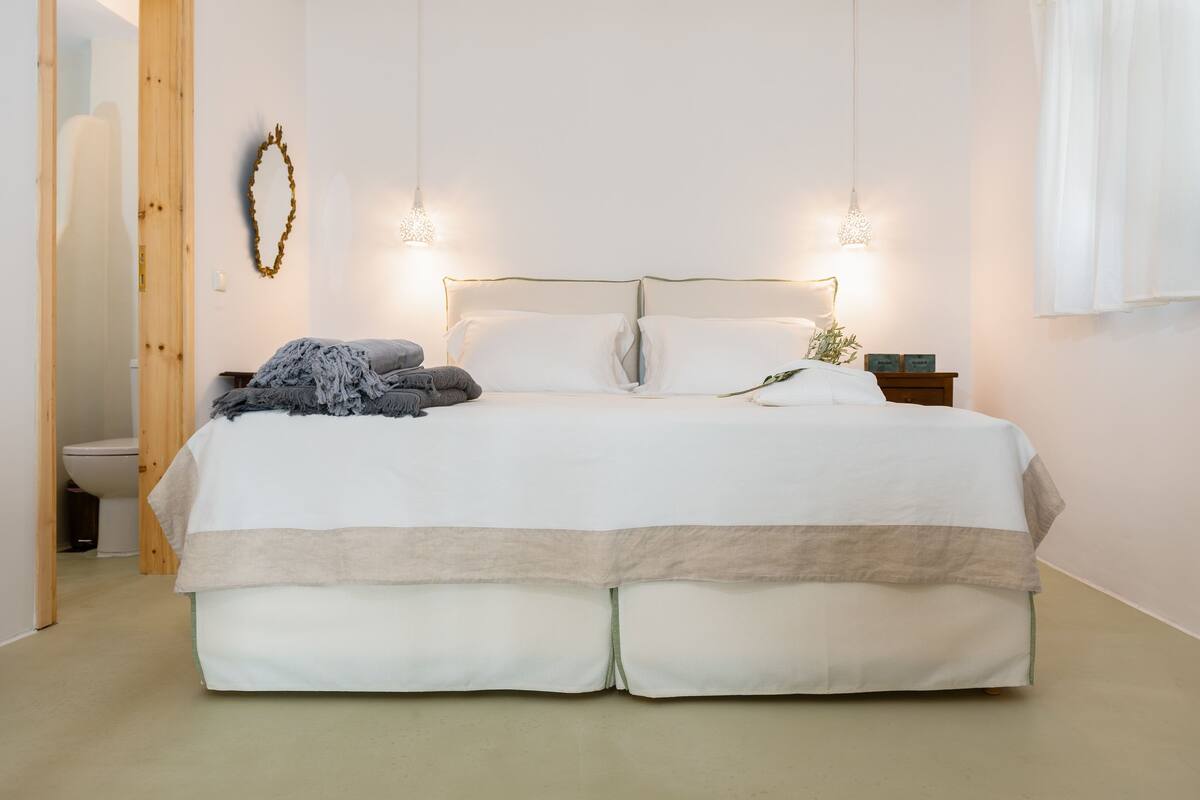 Un lit bien fait, recouvert d’une couvre-lit blanche et beige, avec deux lampes de chevet et une petite table de chevet agrémentée d’une plante.
