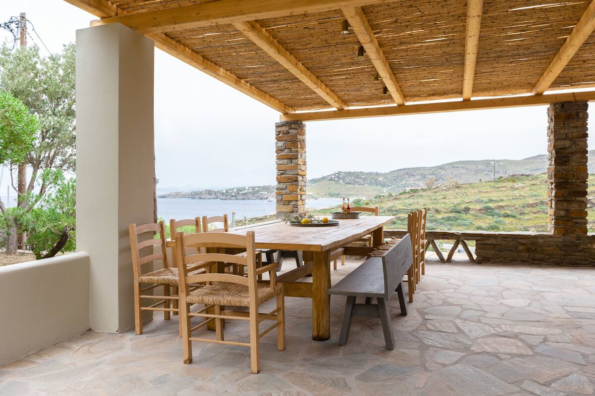 Un espace extérieur abrité, aménagé pour les repas, avec un sol en pierre, des meubles en bois et une vue sur la mer et les collines.