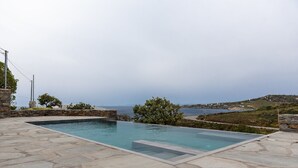 A rectangular swimming pool with stone tiles, surrounded by a stone wall and a view of the sea and hills.