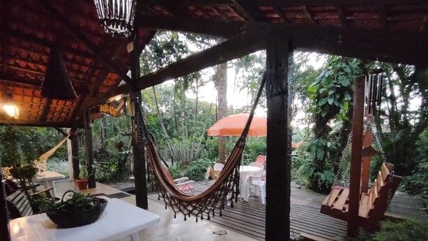 Un espacio al aire libre cubierto con una hamaca, mesas y sillas blancas, rodeado de abundante vegetación.