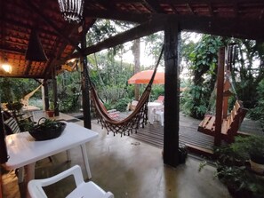 A covered outdoor seating area with a hammock, white tables, and chairs, surrounded by lush greenery. - Pousada Enseada das Conchas (Paranaguá)