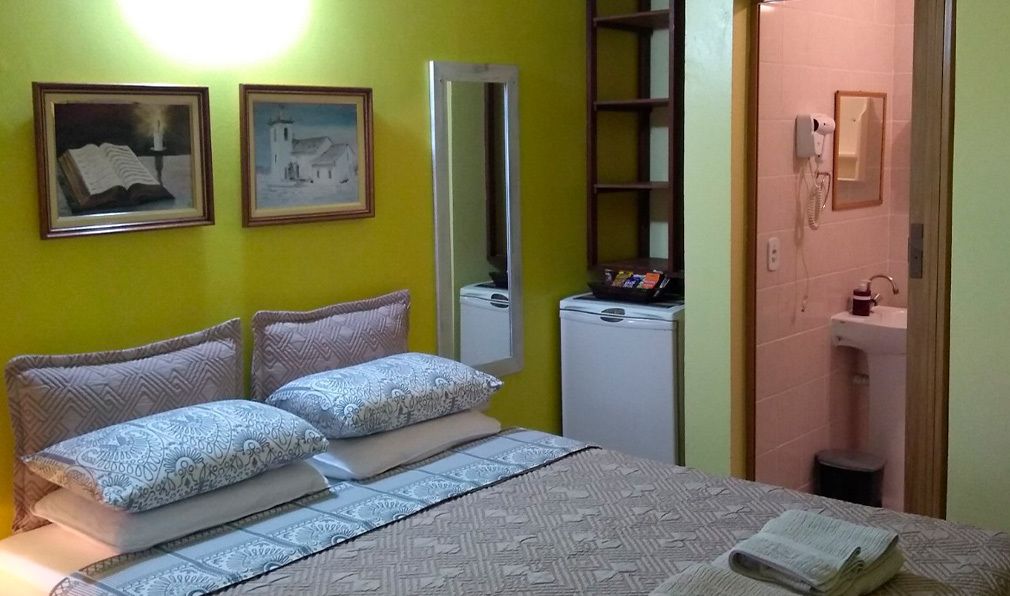 Una habitación con cama, espejo, refrigerador blanco pequeño y baño visible a través de una puerta abierta.