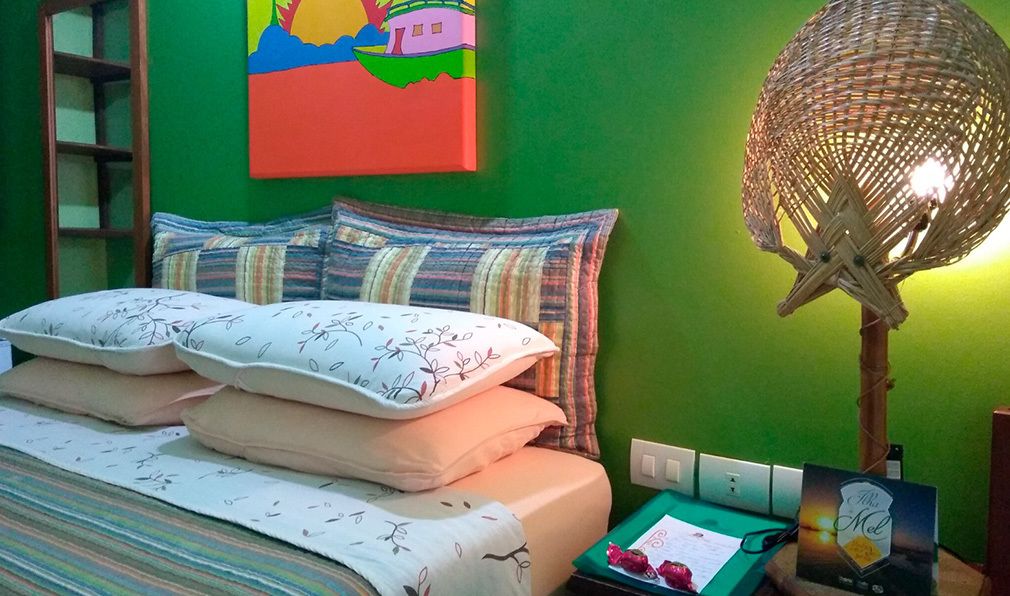 A bed with multiple pillows, a woven lamp, a colorful painting, and a small table with a book and a card.