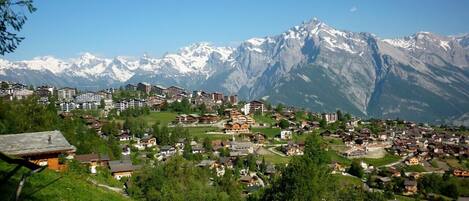 A mountainous village with buildings nestled among the hills, a clear sky, and snow-capped peaks in the background.