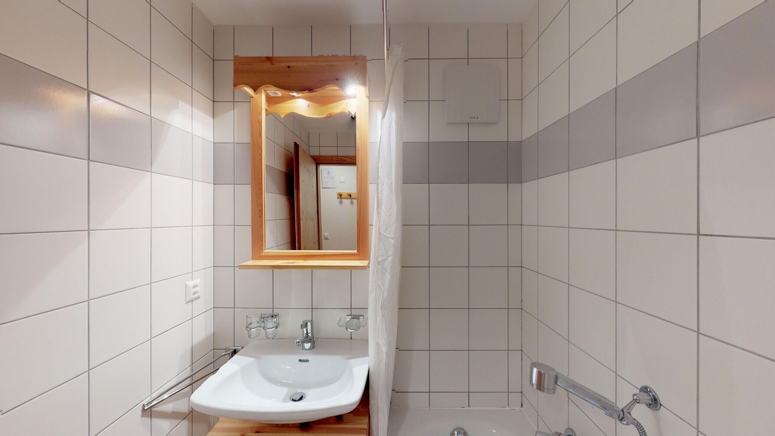 A modern bathroom with white tiles, a wooden-framed mirror, a sink with a faucet, and a shower area.