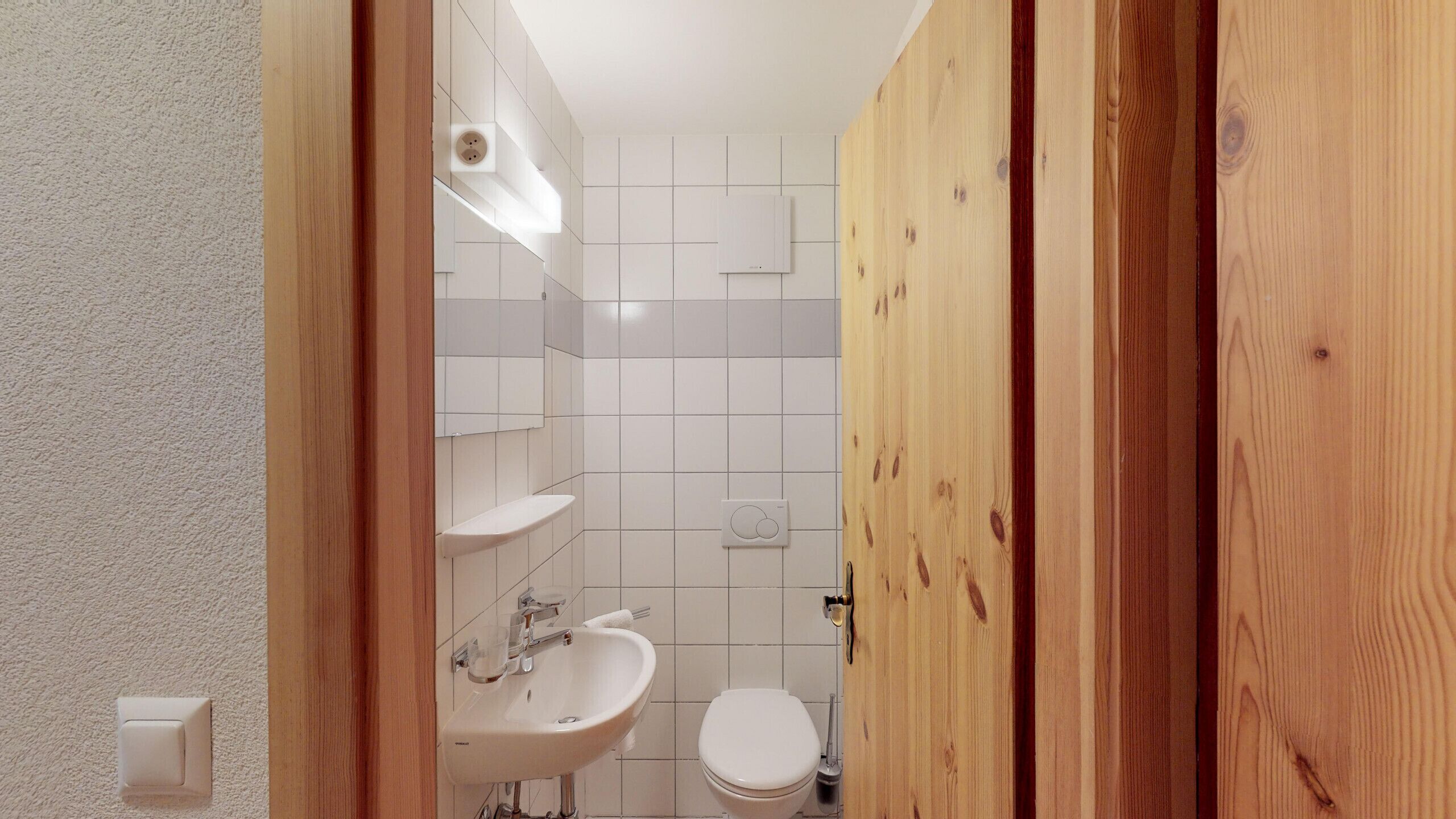 Et lille, moderne badeværelse med hvide fliser, et håndvask, et toilet og en trædør.