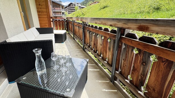 A balcony with wicker furniture and a glass table, overlooking a green hillside with buildings.