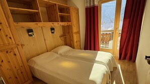 A wooden room with two beds, a wooden wardrobe, and a window with red curtains.