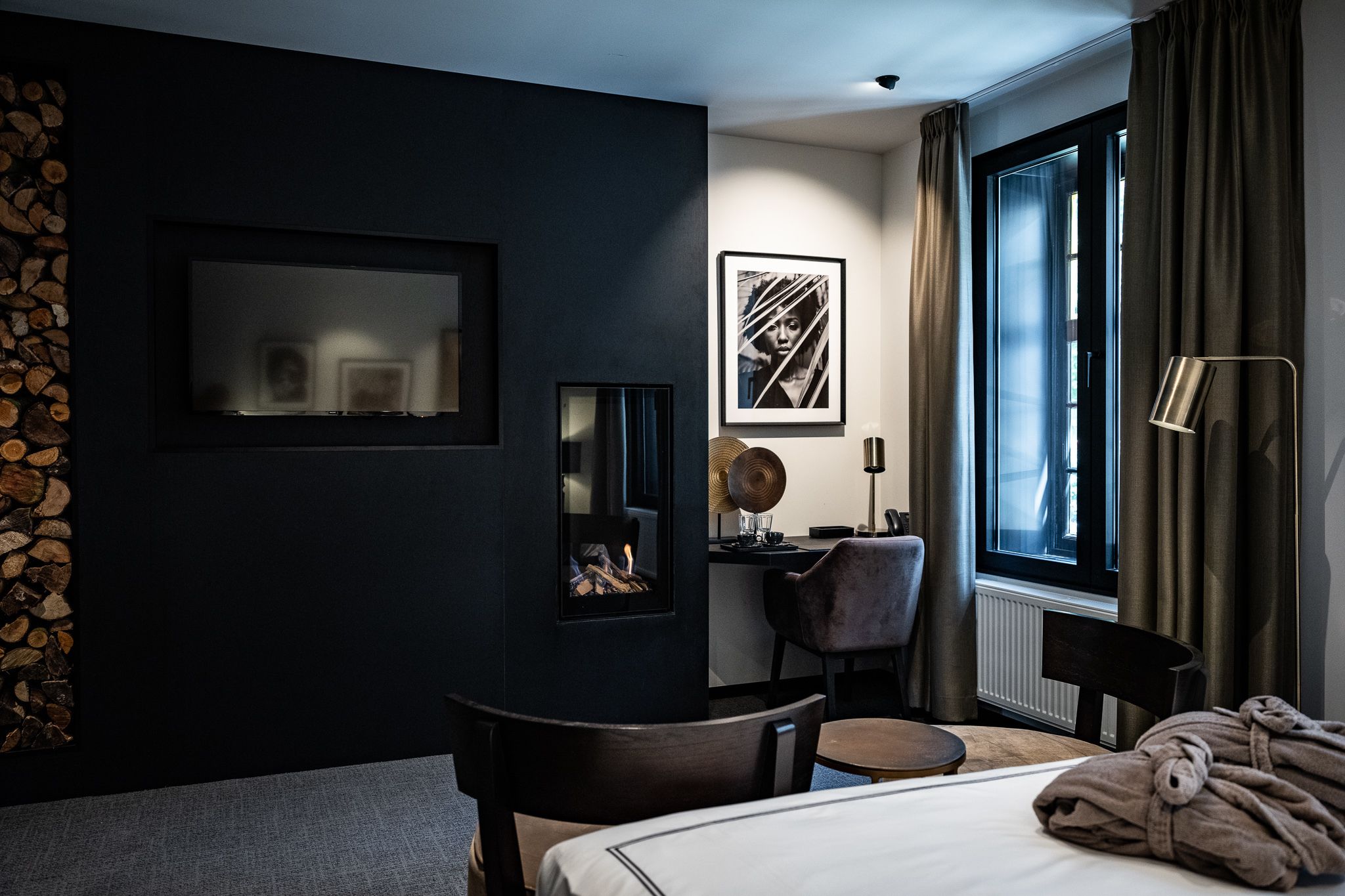 A modern hotel room with a dark wall, a fireplace, a desk, a chair, a bed, and a large window with curtains.