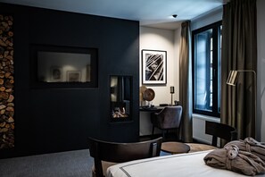Una habitación de hotel moderna con una pared oscura, una chimenea, un escritorio, una silla, una cama y un ventanal grande con cortinas.