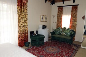 Quarto com uma cama, um pequeno frigorífico branco, um sofá verde com almofadas, um tapete estampado e cortinas com padrão espiralado.