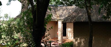 A thatched-roof building with a brick wall, a door, and a window. There is a bench and a table outside.