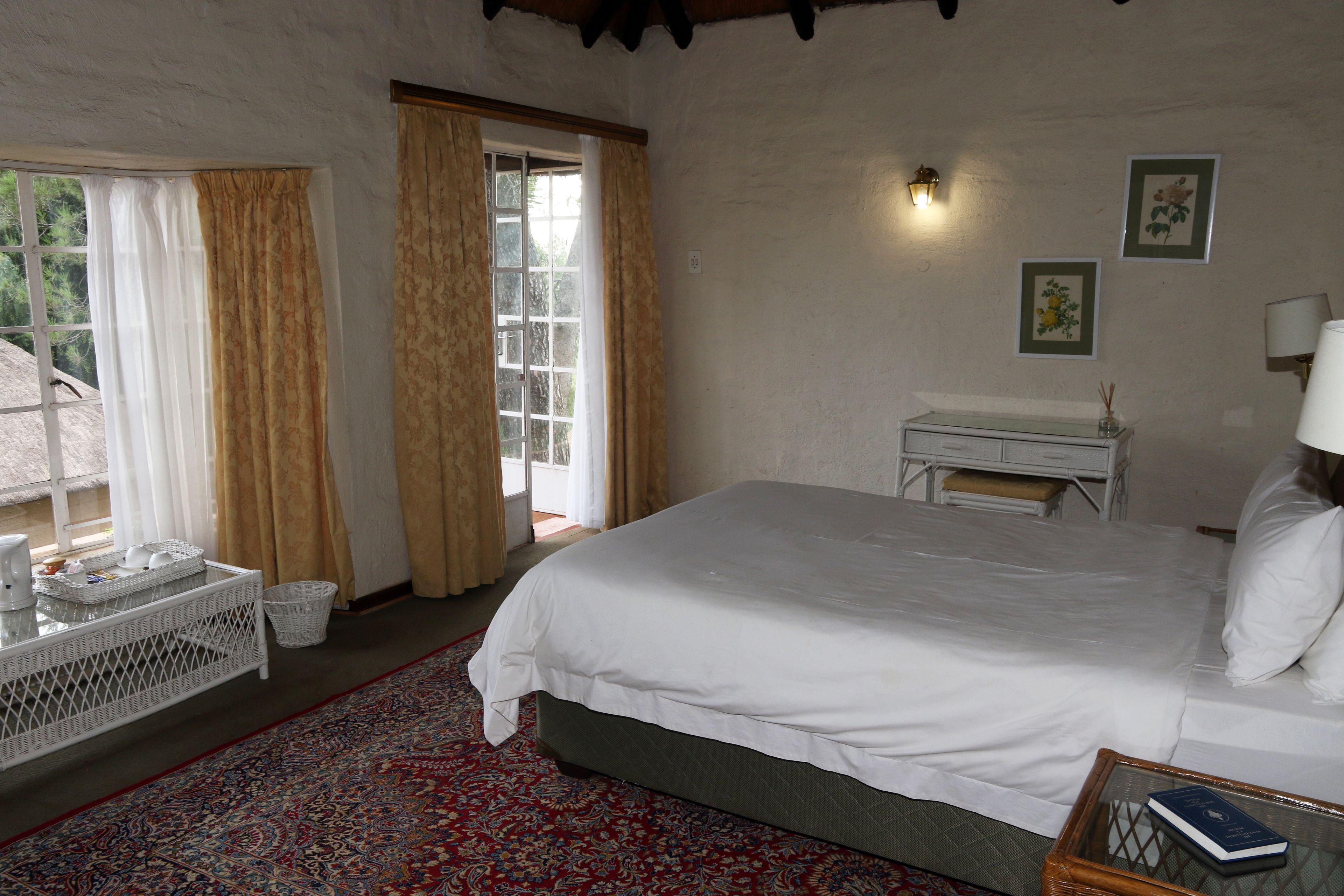 Un dormitorio con una cama grande, una mesita con un libro, una silla de mimbre, una ventana con cortinas y una lámpara de pared.