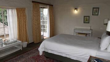 Un dormitorio con una cama grande, una mesita con un libro, una silla de mimbre, una ventana con cortinas y una lámpara de pared.