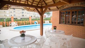 An outdoor pool area with white metal chairs and tables, a brick wall, and a window overlooking the pool. - Elite Hotel (Florianópolis)