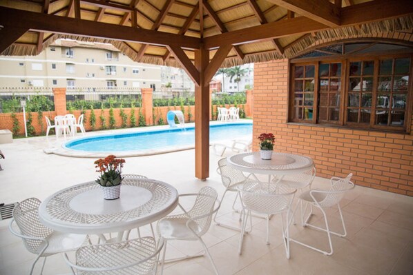 An outdoor pool area with white metal chairs and tables, a brick wall, and a window overlooking the pool. - Elite Hotel (Florianópolis)