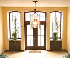 Um corredor com porta de madeira, duas janelas com plantas e um candeeiro pendente.