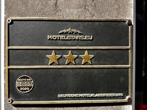 Una placa de hotel con 'HOTELSTAR.S.EU' y tres estrellas doradas, una insignia 'VÁLIDA HASTA: DEHOGA 2029' y el texto 'CLASIFICACIÓN HOTELERA ALEMANA'.