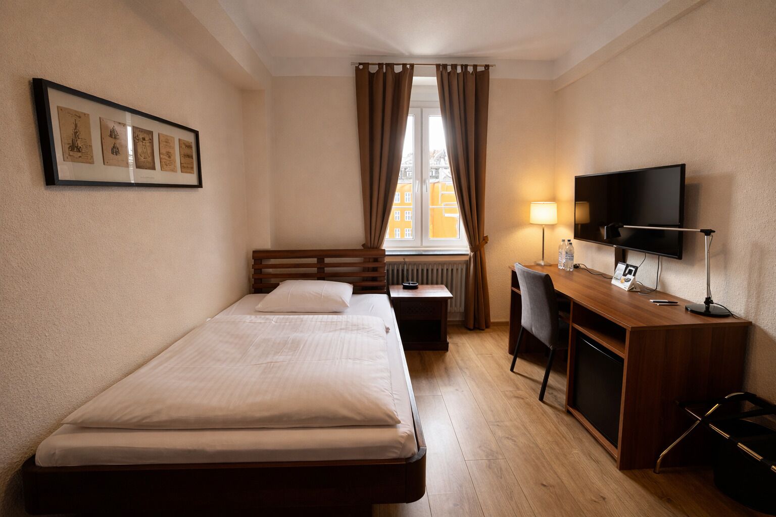 A hotel room with a bed, desk, chair, TV, and framed pictures on the wall.