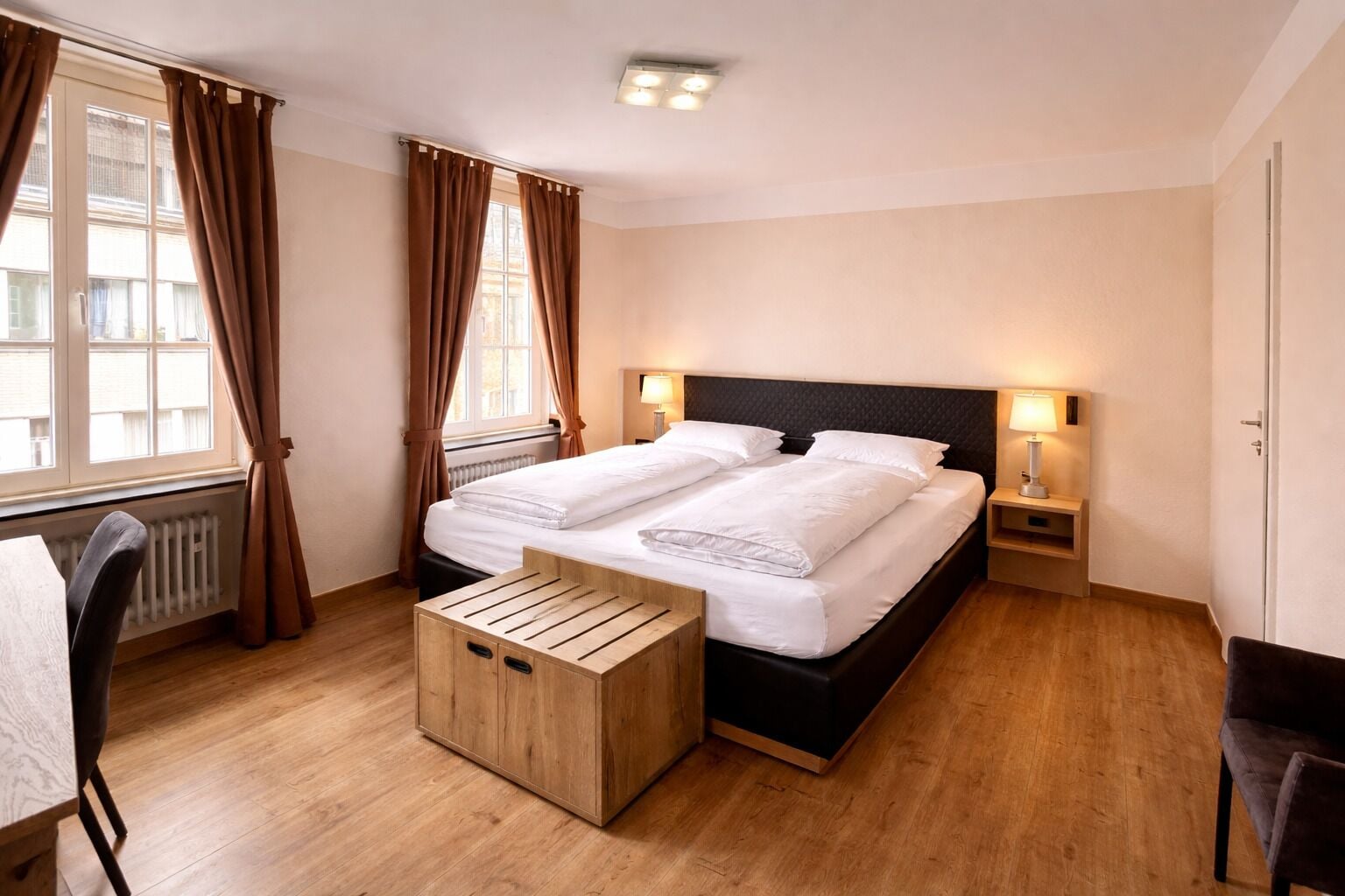 Een hotelkamer met een tweepersoonsbed, nachtlampjes, een houten bankje, een stoel en een radiator.