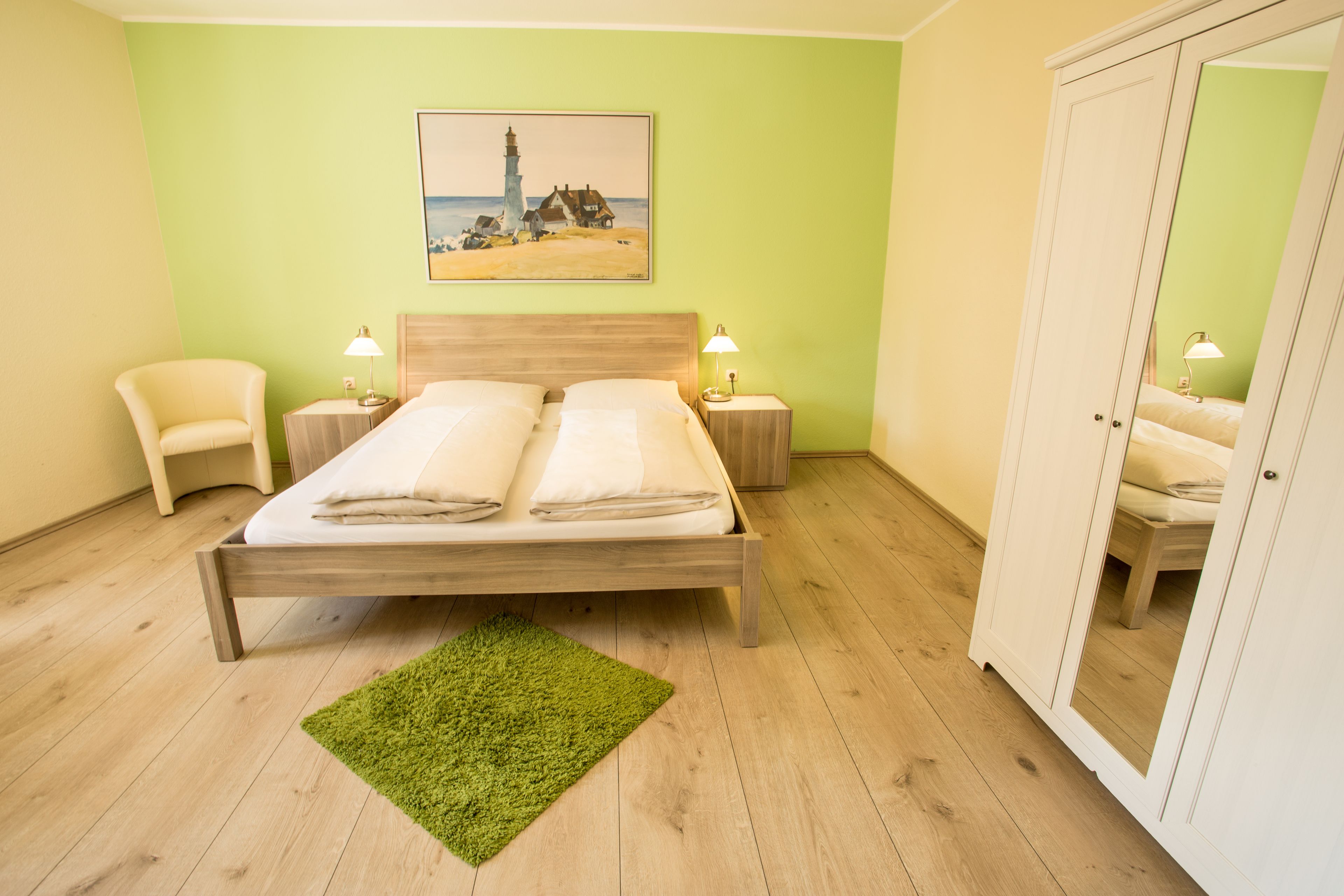 Una cama bien hecha con dos almohadas y una alfombra verde en una habitación con paredes verde claro, suelo de madera y un cuadro de un faro.