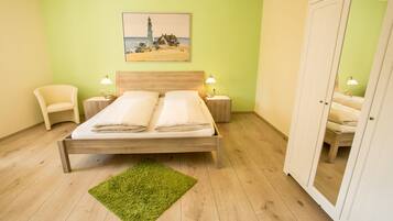 Una cama bien hecha con dos almohadas y una alfombra verde en una habitación con paredes verde claro, suelo de madera y un cuadro de un faro.