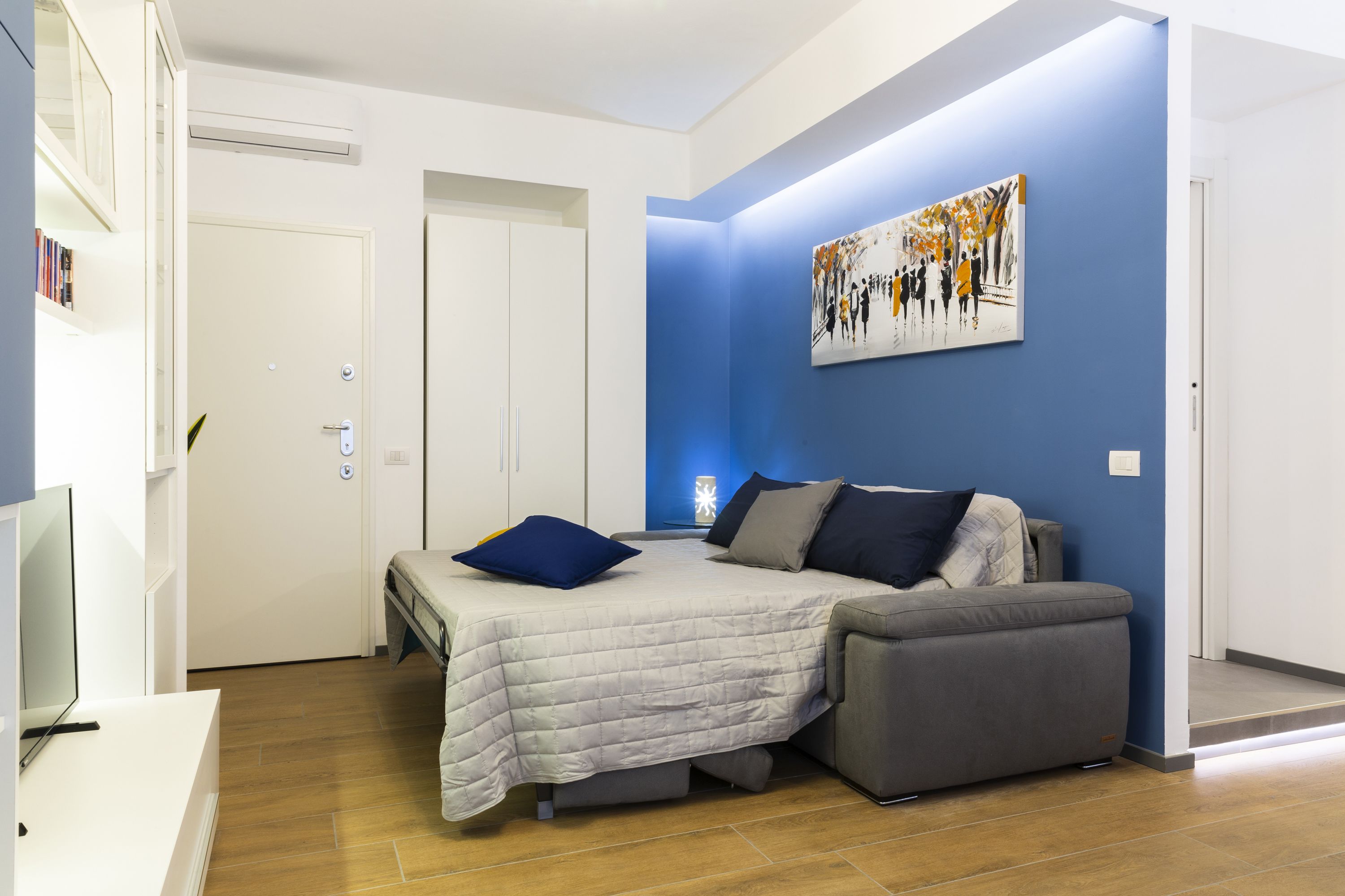 A modern bedroom with a bed, a grey sofa, a painting on the wall, and a television on a stand.