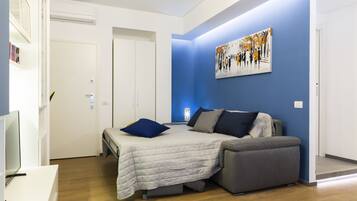 A modern bedroom with a bed, a grey sofa, a painting on the wall, and a television on a stand.