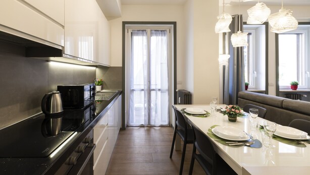 Una cucina moderna con piano di lavoro nero, elettrodomestici in acciaio inossidabile e zona pranzo con tavolo bianco apparecchiato per due.