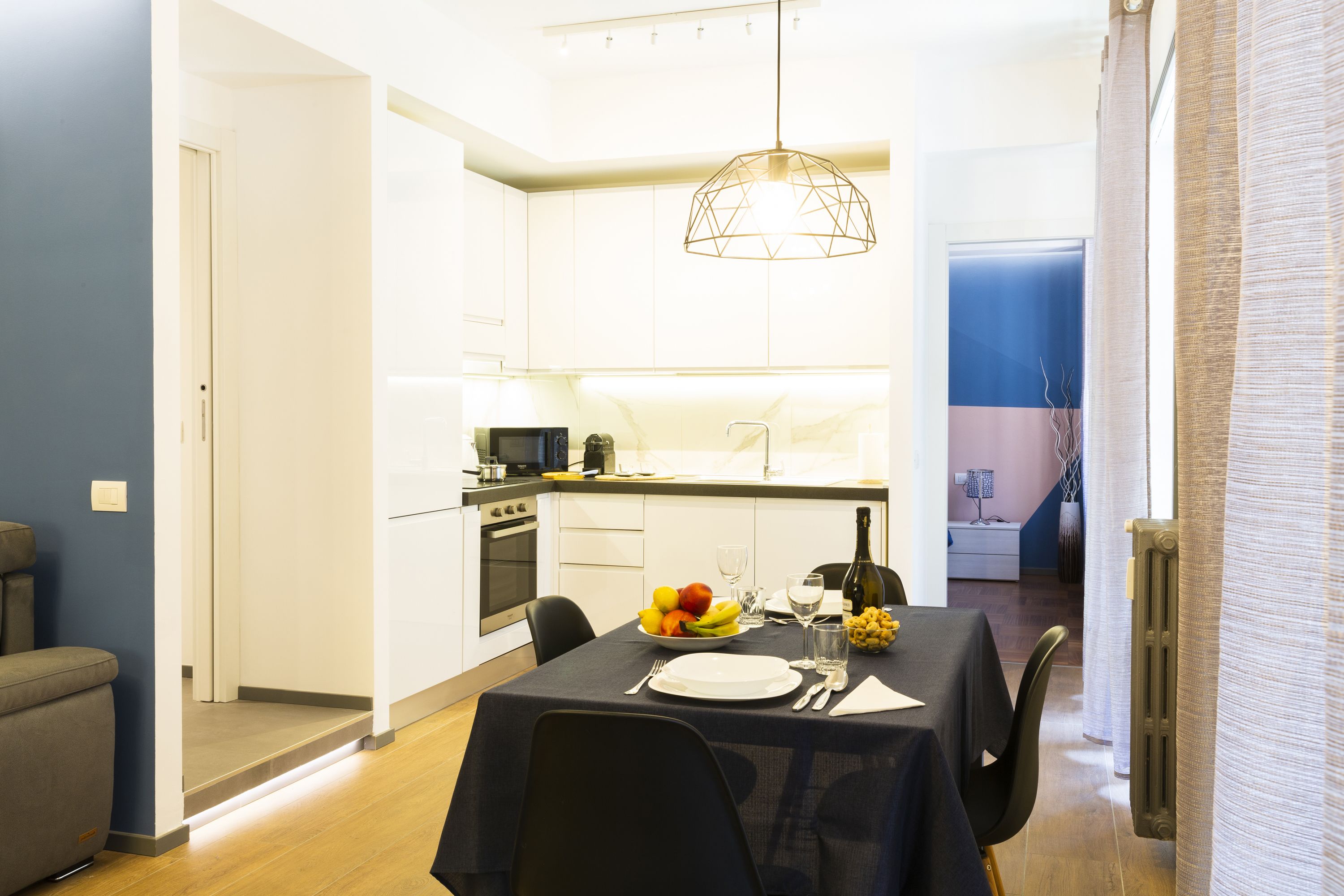 Una cocina moderna con armarios blancos, un comedor con una mesa dispuesta para dos personas y una lámpara colgante.