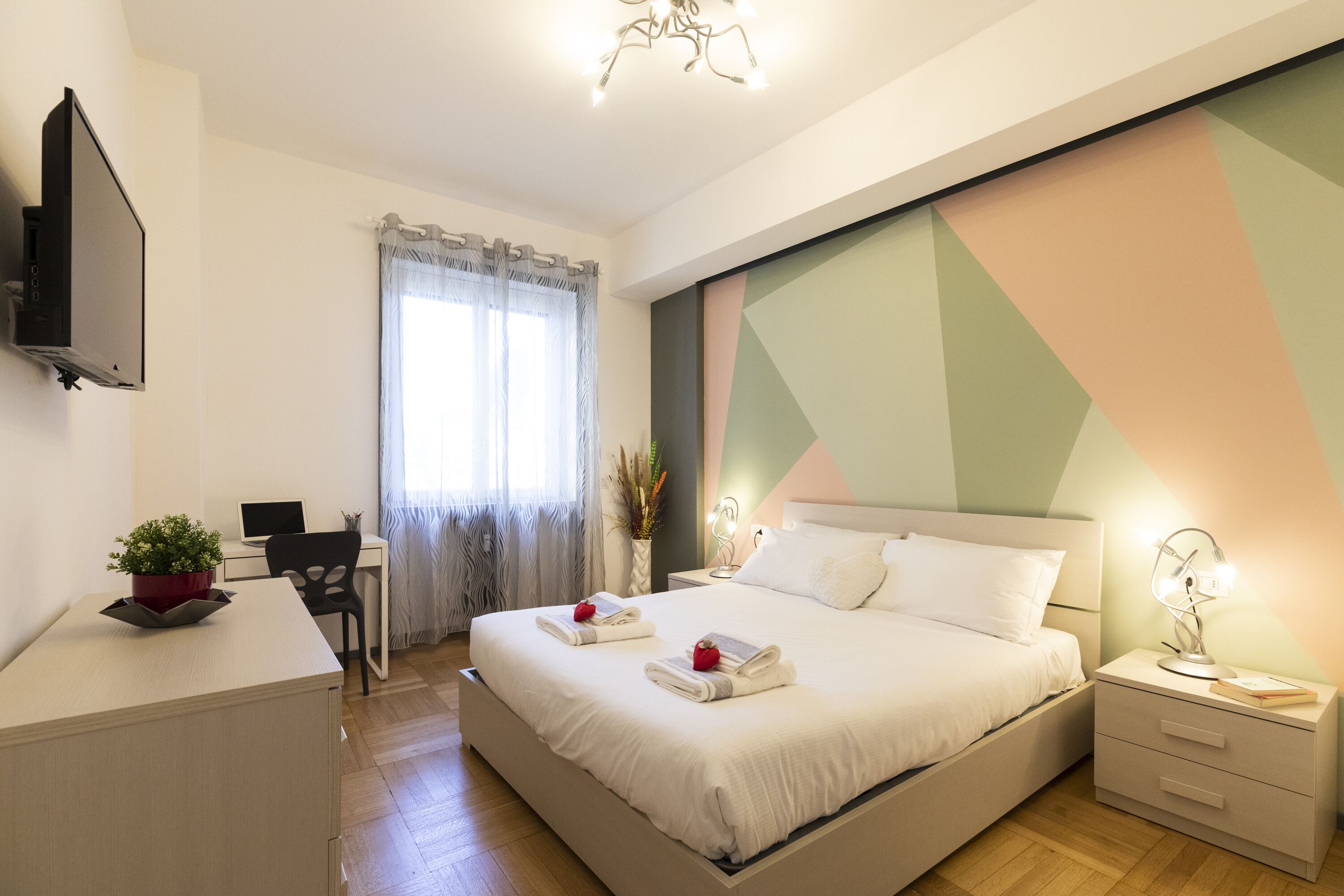 A modern bedroom with a bed, bedside table, desk, chair, and a TV mounted on the wall.