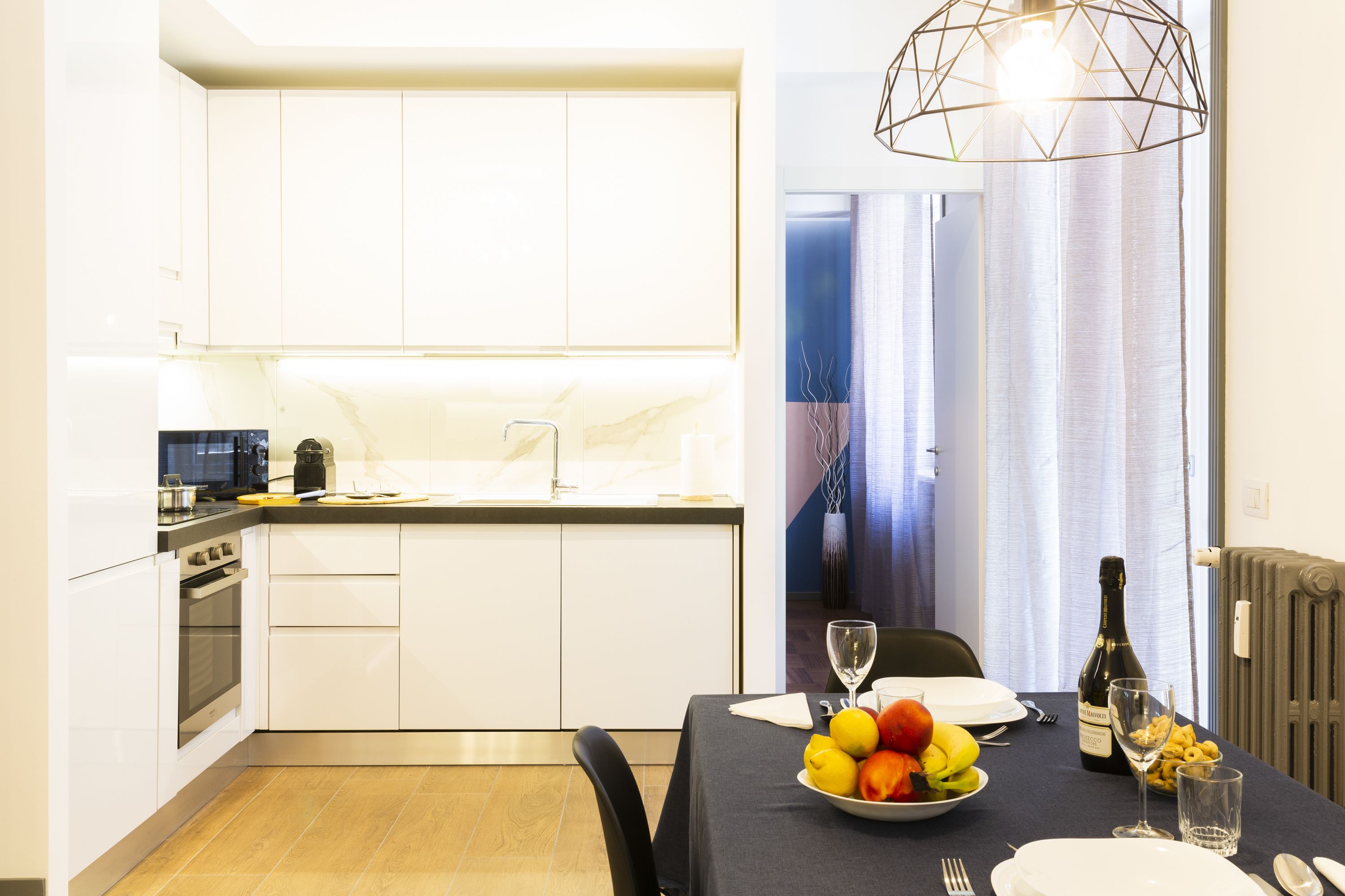 Une cuisine moderne avec des meubles blancs, une table à manger dressée avec des assiettes, un saladier rempli de fruits et une bouteille de champagne.