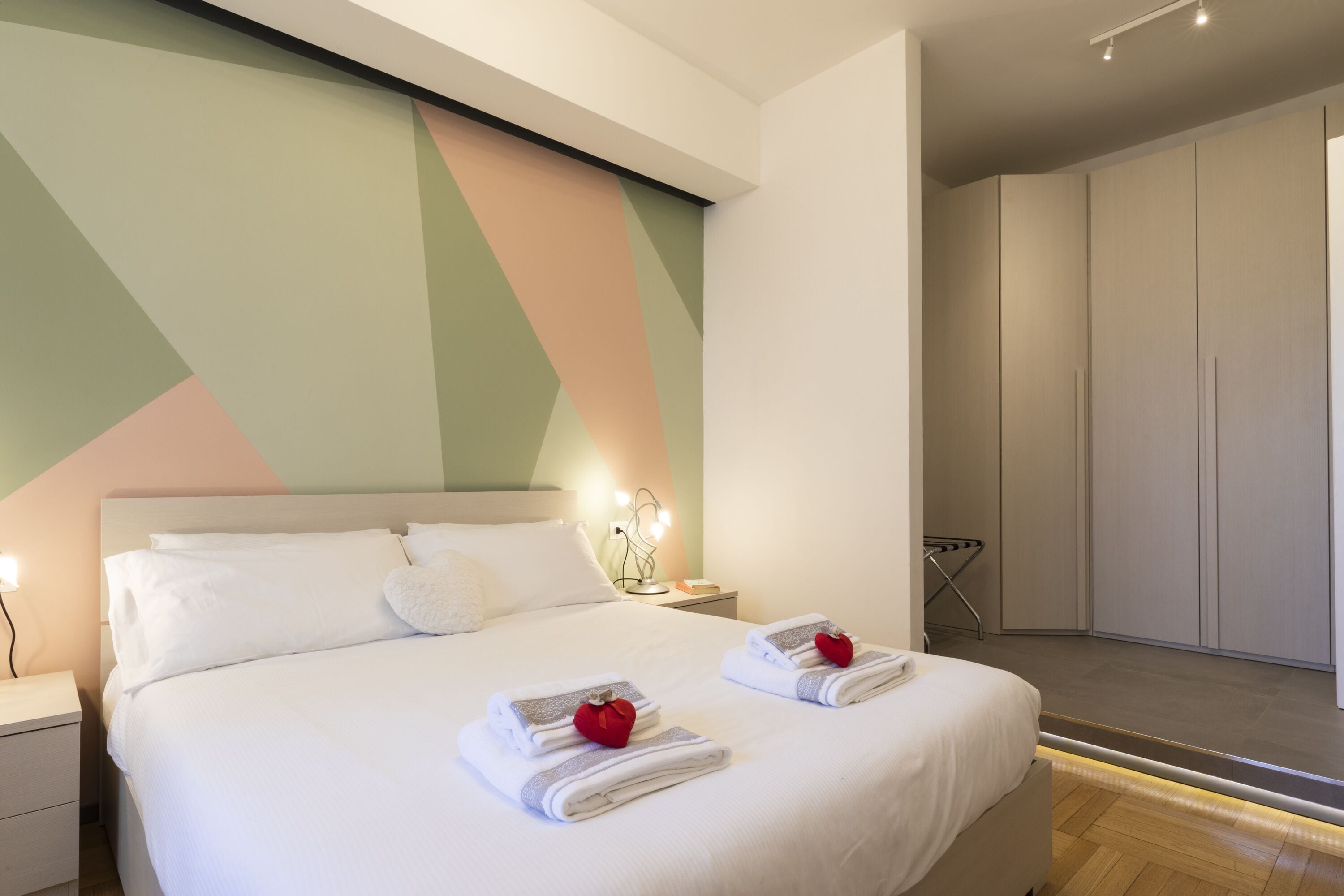 A modern hotel room with a large bed, a bedside table with a lamp, and a wardrobe.