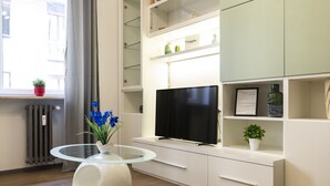 Un soggiorno moderno con un tavolino da caffè in vetro, una televisione a schermo piatto, scaffalature a muro e una pianta in vaso.