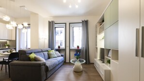 Un soggiorno moderno con un divano grigio, un tavolino in vetro e un angolo cottura con mobili bianchi e un frigorifero.